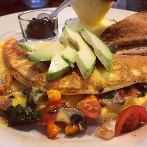A delicious omelette filled with vegetables, topped with avocado slices, served with toast and a side of jam.