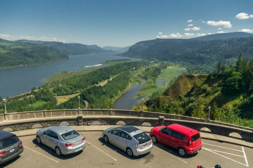 A scenic view of a river valley surrounded by mountains, with parked cars in the foreground under a clear blue sky.