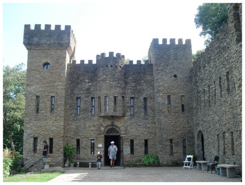A stone castle with towers and arched entrance, surrounded by greenery and people walking nearby.