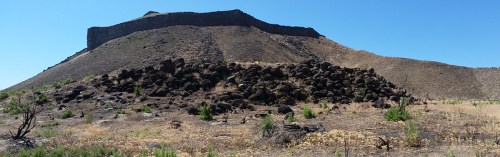 A panoramic view of a rocky hill with a flat top, surrounded by dry grass and sparse vegetation under a clear blue sky.