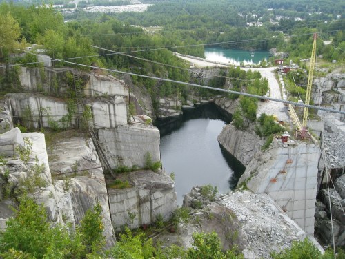 Aerial view of a stone quarry with a deep water-filled pit surrounded by rocky cliffs and greenery.