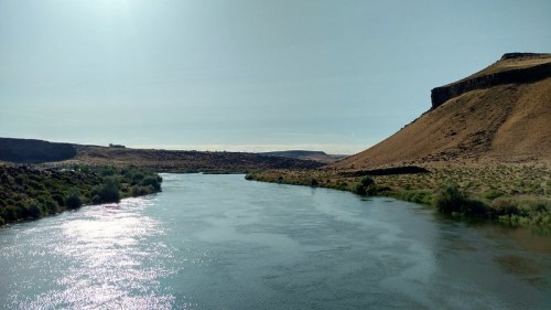 A serene river flows through a landscape of hills and greenery under a clear blue sky. Sunlight sparkles on the water.