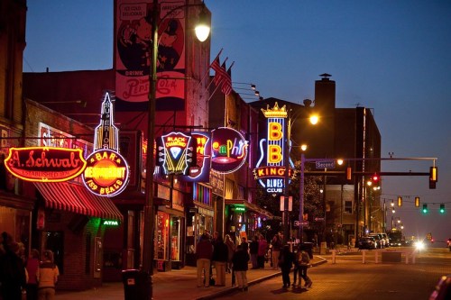 Vibrant street scene at dusk with neon signs for bars and clubs, people walking, and traffic lights in a lively urban setting.
