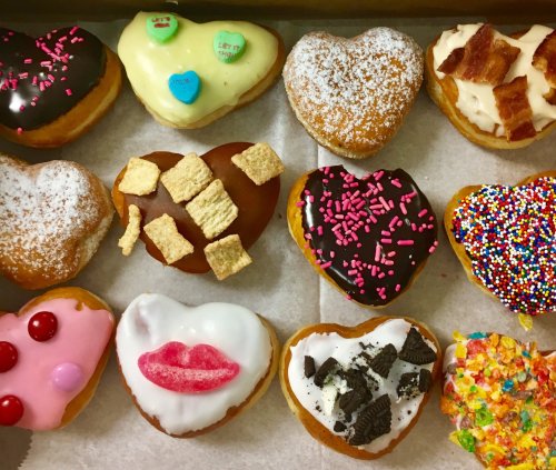 A variety of heart-shaped donuts with colorful toppings, including sprinkles, frosting, and cereal, arranged in a box.