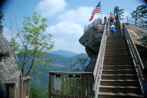 A wooden staircase leads to a viewpoint with an American flag, surrounded by mountains and trees.