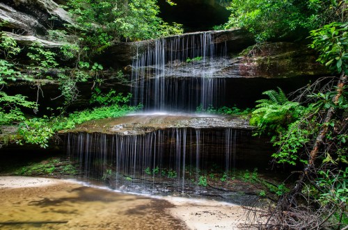 A serene waterfall cascades over rocky ledges, surrounded by lush greenery and a calm pool below.