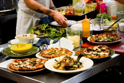 A busy kitchen counter with various pizzas, a salad, and colorful ingredients in jars, as a chef prepares food.