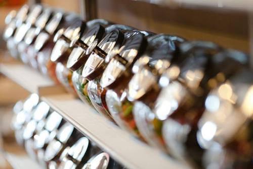 A row of glass jars with metal lids, filled with colorful ingredients, displayed on a shelf.