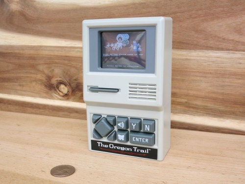 A vintage handheld game console displaying "The Oregon Trail" on its screen, with buttons and a coin for scale.