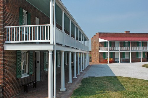 Historic brick buildings with white columns and green shutters, surrounding a grassy courtyard under a clear blue sky.