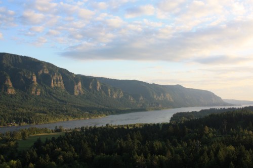 A scenic view of a river winding through lush green forests and mountains under a partly cloudy sky.