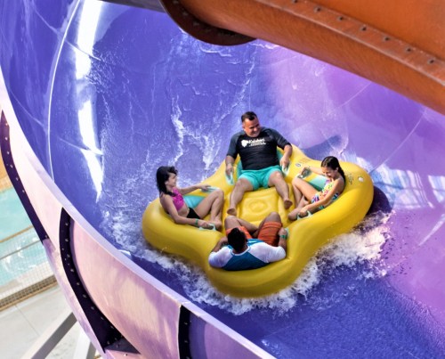 A group of four people enjoying a water slide in a colorful, twisting tube, splashing through the water.
