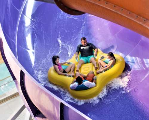 A group of four people enjoying a water slide in a colorful, twisting tube, splashing through the water.