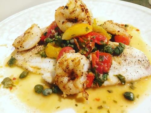 A plate of seasoned fish topped with shrimp, cherry tomatoes, and herbs in a flavorful sauce.