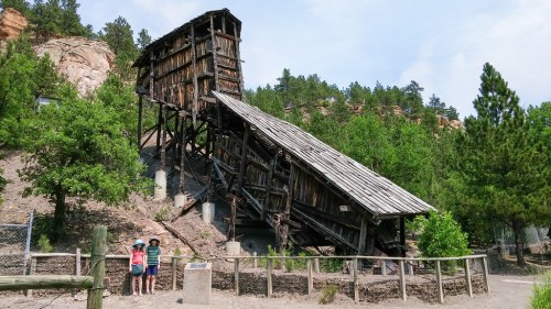 Two children stand near a large, tilted wooden structure surrounded by trees and rocky terrain.