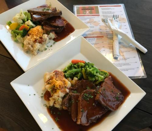 Two plates of hearty meals featuring beef, gravy, mashed potatoes, and mixed vegetables, with a menu in the background.