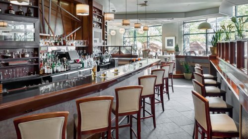A modern bar area with a sleek counter, high stools, and a well-stocked shelf of liquor, surrounded by large windows.