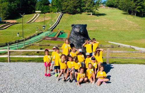 A group of children in yellow shirts poses in front of a large bear statue at an outdoor activity area.