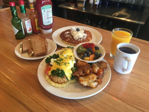 A breakfast spread featuring eggs Benedict, pancakes, fruit, toast, coffee, and orange juice on a wooden table.