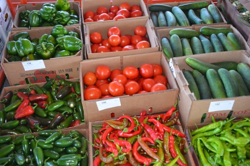 Colorful assortment of fresh vegetables in boxes, including green peppers, tomatoes, cucumbers, and various chili peppers.