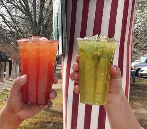 Two hands hold colorful drinks in clear cups: one red and one green, both filled with ice.