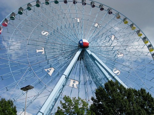 A blue Ferris wheel with colorful gondolas and a Texas flag at the center, set against a cloudy sky.