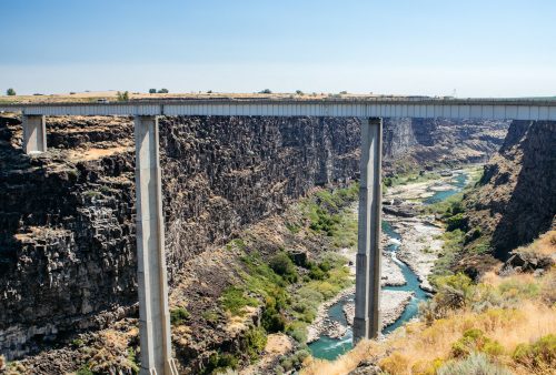 A concrete bridge spans a deep canyon with a river below, surrounded by rocky cliffs and sparse vegetation under a clear sky.