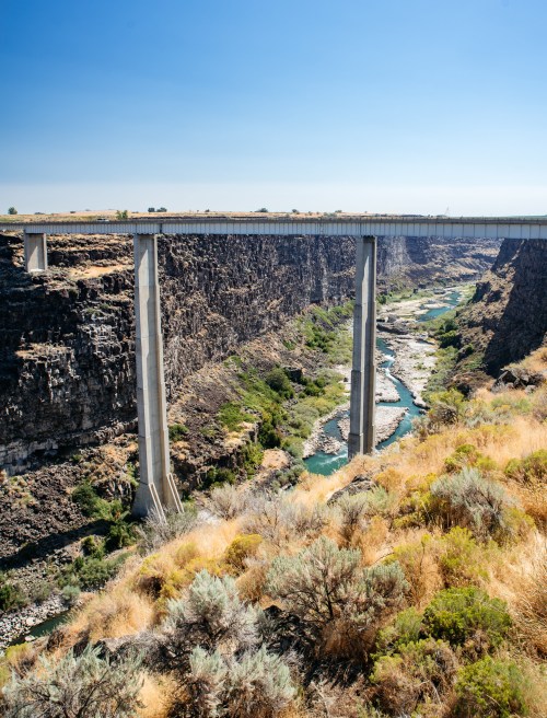 A tall bridge spans a deep canyon with a river below, surrounded by rocky cliffs and sparse vegetation under a clear blue sky.