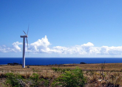 A wind turbine stands on a grassy hillside overlooking the ocean under a clear blue sky with fluffy white clouds.