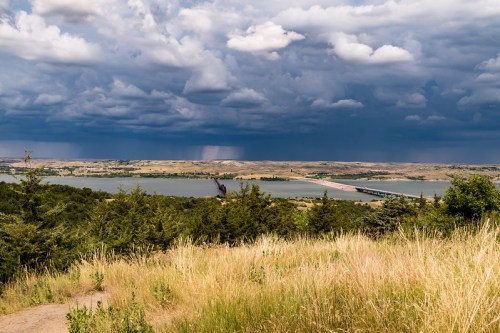 A scenic view of a river and bridge under a dramatic sky filled with dark clouds and distant rain. Lush greenery in the foreground.