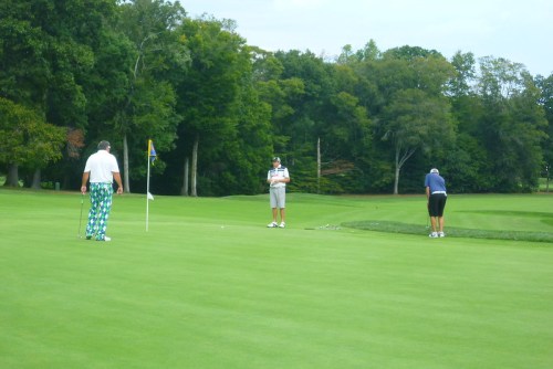 Three golfers on a green, one putting near the hole, surrounded by trees and a clear sky.
