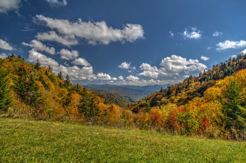 A vibrant autumn landscape with colorful trees, rolling hills, and a clear blue sky dotted with fluffy clouds.