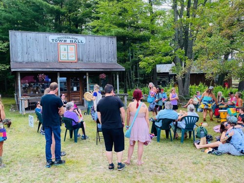 A lively outdoor gathering at Michigamme Town Hall, with people playing drums and enjoying music in a forested setting.