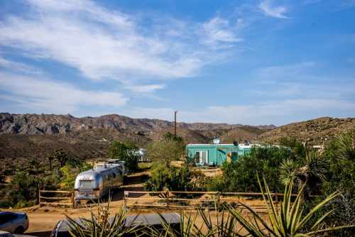 A desert landscape featuring a turquoise house, an Airstream trailer, and mountains under a blue sky.