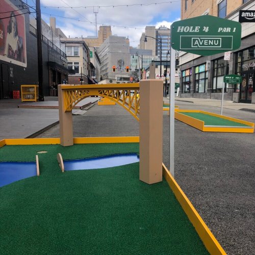 Mini golf course featuring a bridge and a sign for "Hole 24, Par 1" in an urban setting.