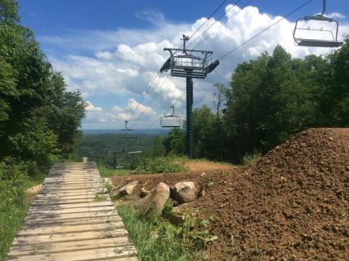 A scenic view of a ski lift with empty chairs, surrounded by trees and a dirt mound, under a blue sky with clouds.