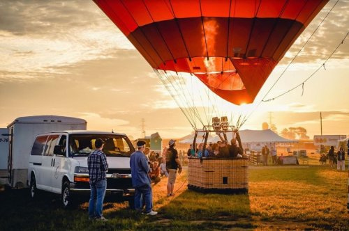 A hot air balloon prepares for takeoff at sunrise, with people gathered around and a van nearby.