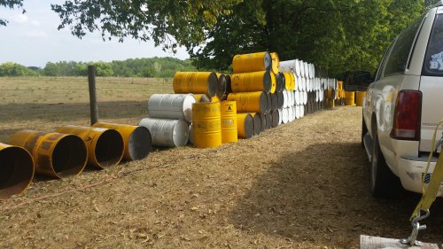A row of stacked yellow and white barrels beside a vehicle in a grassy field with trees in the background.