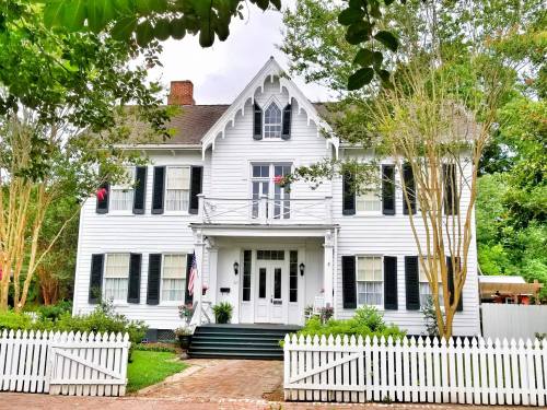 A charming white two-story house with black shutters, a front porch, and a white picket fence, surrounded by greenery.