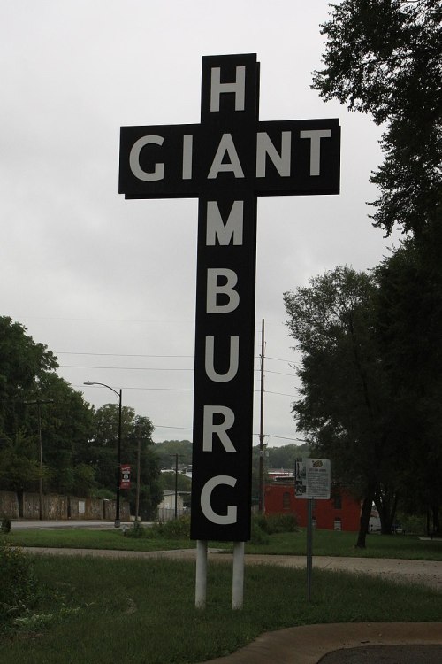 A large black and white sign reading "GIANT HAMBURG" stands on a roadside, surrounded by greenery and overcast skies.