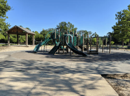A playground with slides and climbing structures, surrounded by trees and a picnic area under a clear blue sky.
