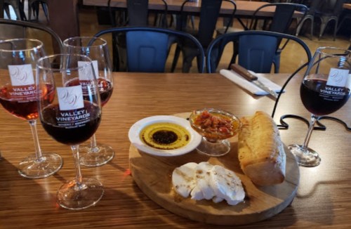 A wooden platter with bread, cheese, and dips, accompanied by four glasses of wine on a table.
