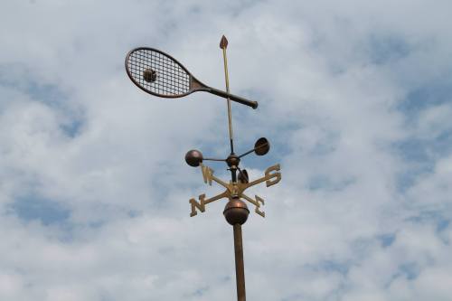 A whimsical weather vane shaped like a tennis racket, with directional indicators, against a cloudy sky.