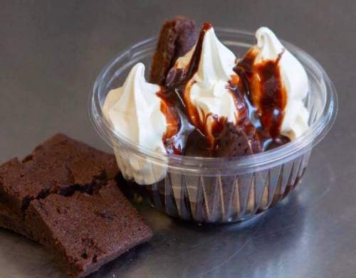 A clear cup filled with whipped cream, brownie pieces, and chocolate sauce, alongside a brownie square.