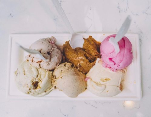 A platter of six ice cream scoops in various flavors, with two spoons resting on top.