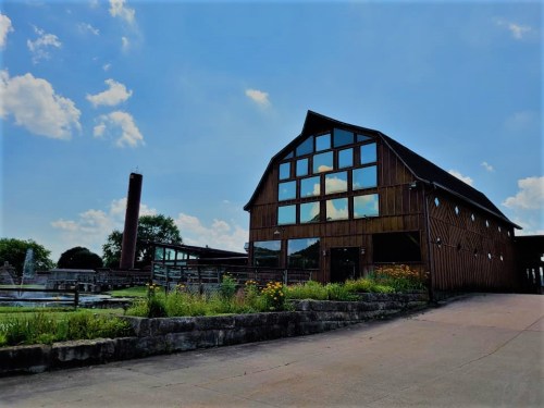 A large wooden barn with large windows, surrounded by greenery and a clear blue sky with scattered clouds.