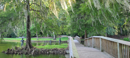 A wooden bridge leads to a serene pond surrounded by trees and sculptures of figures near the water's edge.