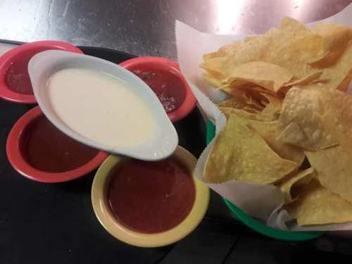 A basket of tortilla chips with three bowls of dipping sauces: two red salsas and one creamy dip.