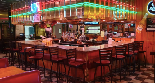 A cozy bar with a wooden counter, colorful neon lights, and empty stools in a casual dining setting.