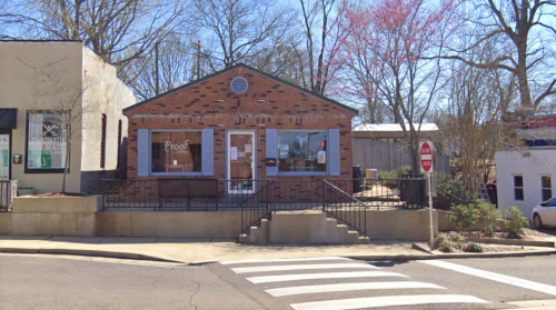 A brick building with large windows, labeled "Proof," situated on a street corner with trees and a stop sign nearby.